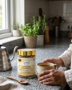 Person holding a cup of tea next to a jar of Deshla Arjuna Plus Herbal Infusion on a kitchen counter.