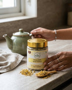 Person holding a jar of chamomile dried flowers on a kitchen counter with a teapot and cloth in the background.