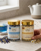 Two jars of Deshla tea on a table with a hand holding one jar, surrounded by tea leaves and a teapot.