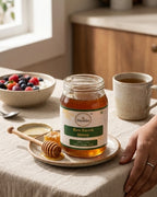 Jar of Desha Raw Forest Honey on a table with a bowl of berries and a cup of tea.